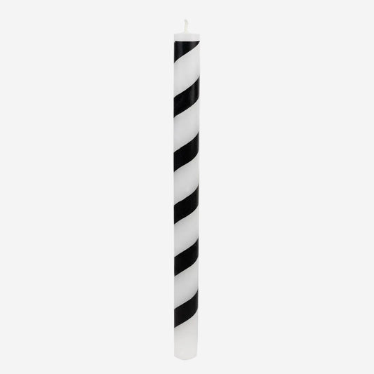 A tall, slender taper candle with alternating diagonal black and white stripes, standing upright against a solid white background.