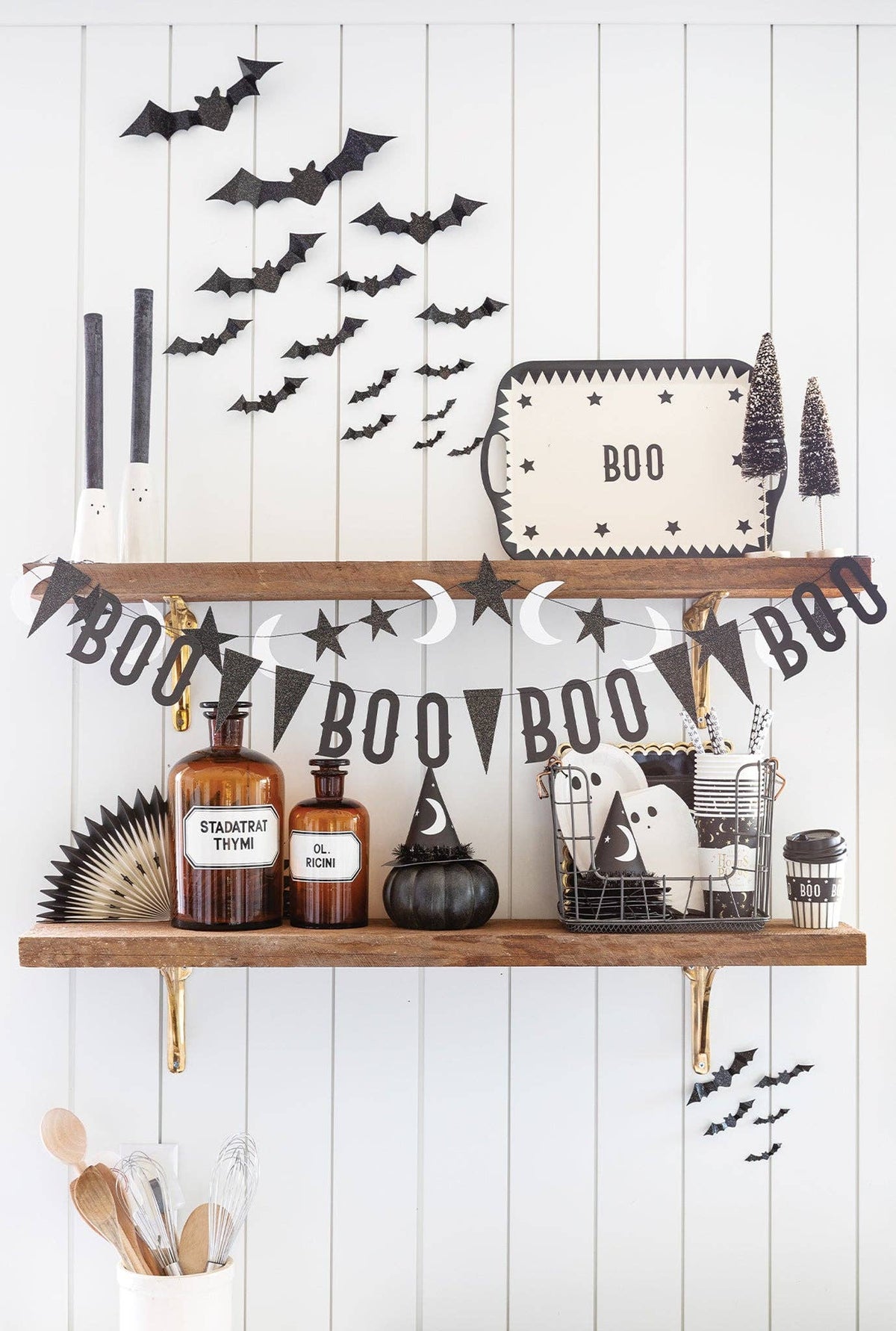 Halloween-themed shelf decor arrangement on two wooden floating shelves against a white shiplap wall, featuring black bat cutouts, a 'BOO' banner, star and moon garlands, amber bottles, a small black pumpkin, a tray sign, and spooky accessories.