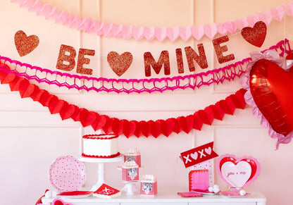 Decorative setup with 'Be Mine' banner, balloons, and Valentine's Day-themed items on a light pink background.